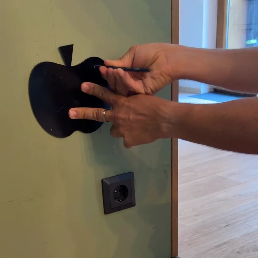 Zwei Hände halten die ausgeschnittene Apfelplatte an die Wand und zeichnen mit einem Stift durch eines der Schraublöcher an