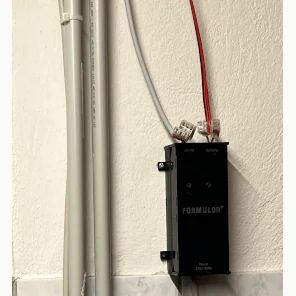 Der Kasten hängt an zwei schwarzen Winkeln an der weiß gestrichenen Wand des Heizungsraums, daneben laufen Leerrohre, von oben kommen ein rotes und ein graues Kabel in Klemmen
