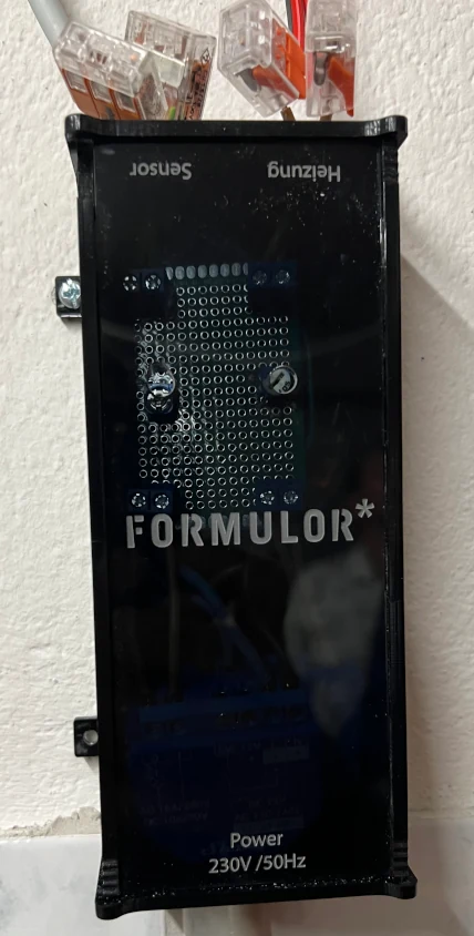 Der fertige Manipulator an der Wand: ein flacher Kasten aus dunklem, durchscheinendem Acrylglas, durch die Front schimmern Lochrasterplatine und Funkrelais; eingraviert die Wortmarke FORMULOR*, oben die Beschriftungen Sensor und Heizung, unten Power 230V /50Hz