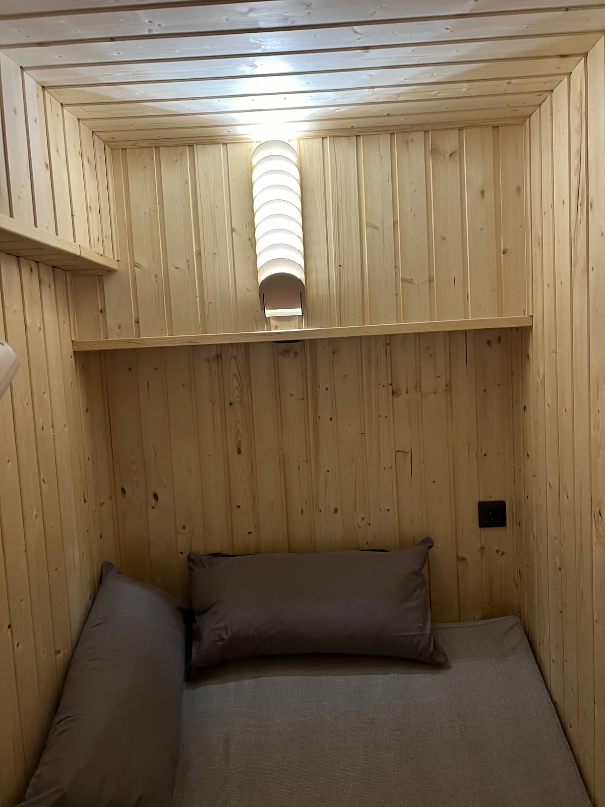 Holzverkleidete Schlafnische mit Matratze und Kissen, an der Wand darüber die eingeschaltete Lamellenlampe als einzige Lichtquelle